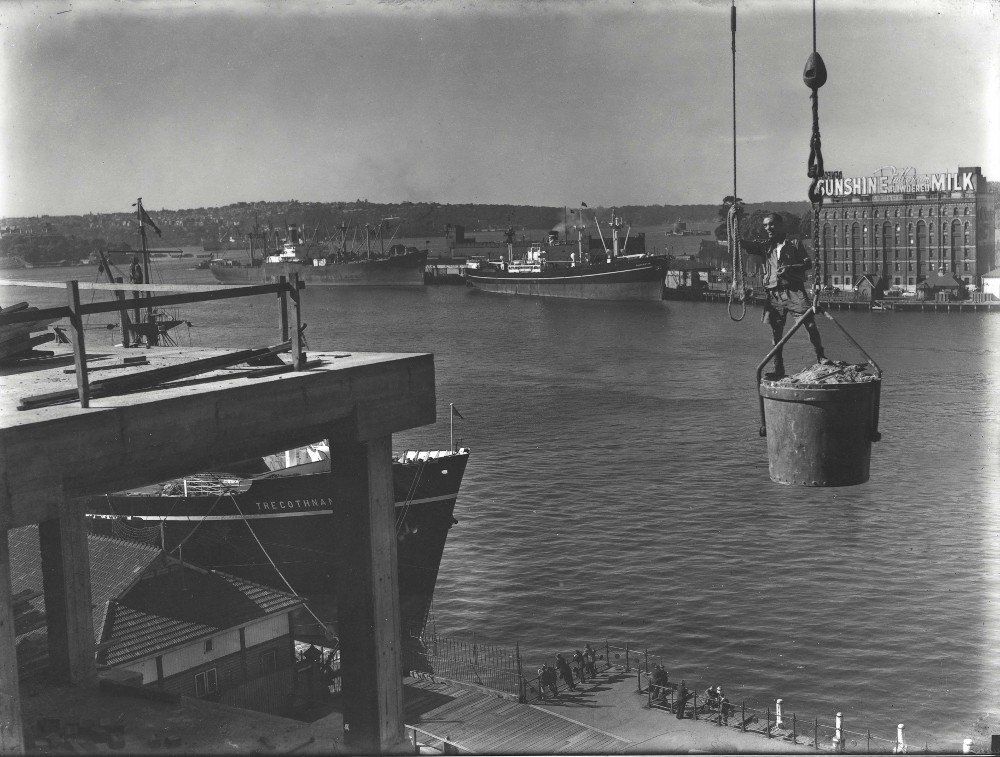 View of shipping wharves in Sydney Harbour with a construction worker standing in the bucket of a crane in the foreground (c. 1959)