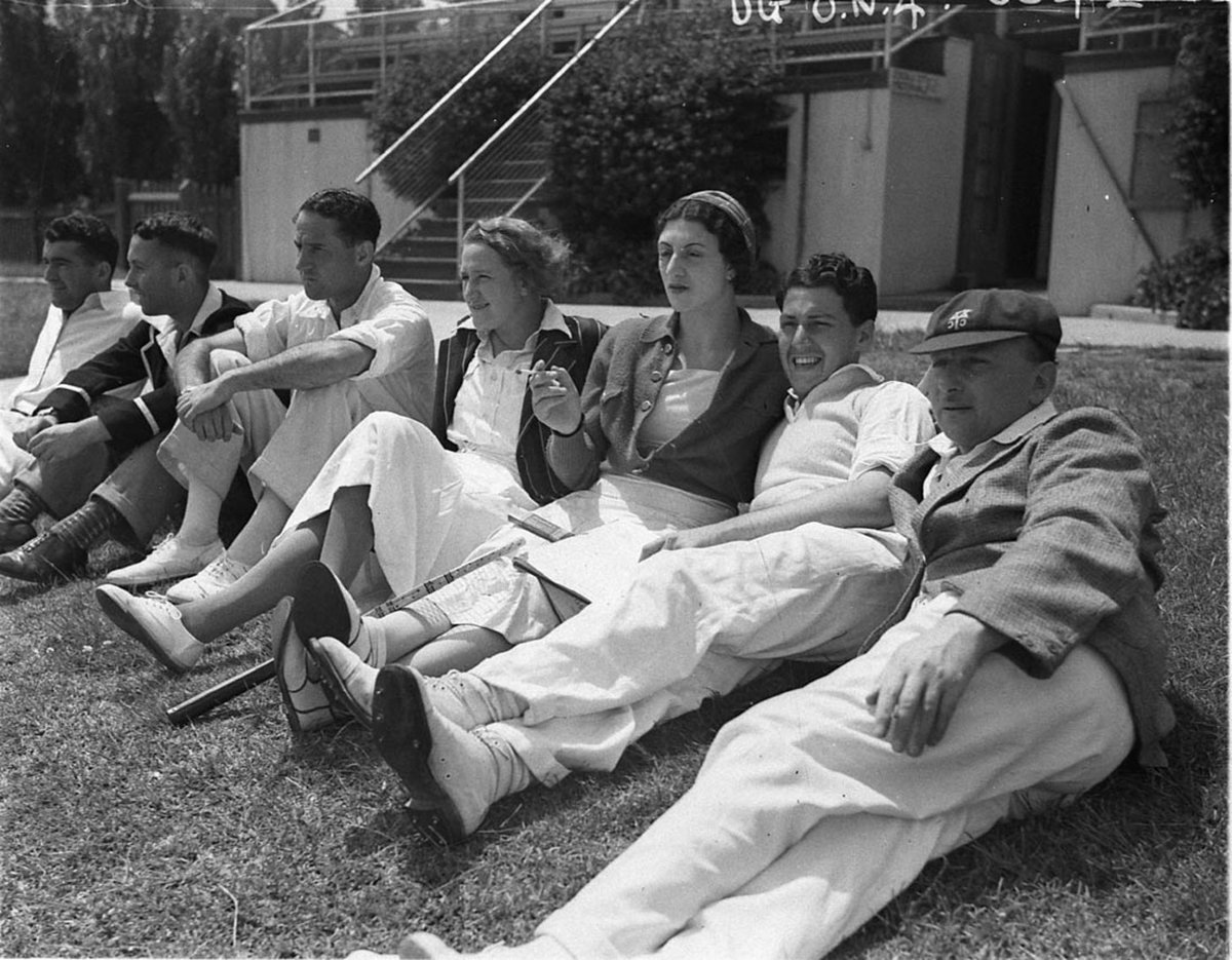 Cricketers and some female spectators relax on the grass bank (1930s)