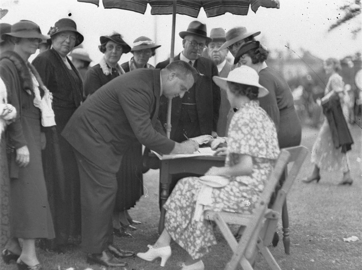 NSW Premier BSB Stevens signs something for a woman sitting under a sunumbrella (1935?)