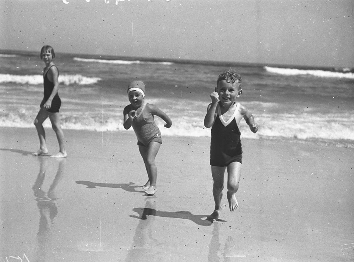 Two children photographed with wrestler Lurich on the same day run up the sand Bondi Beach (1935)