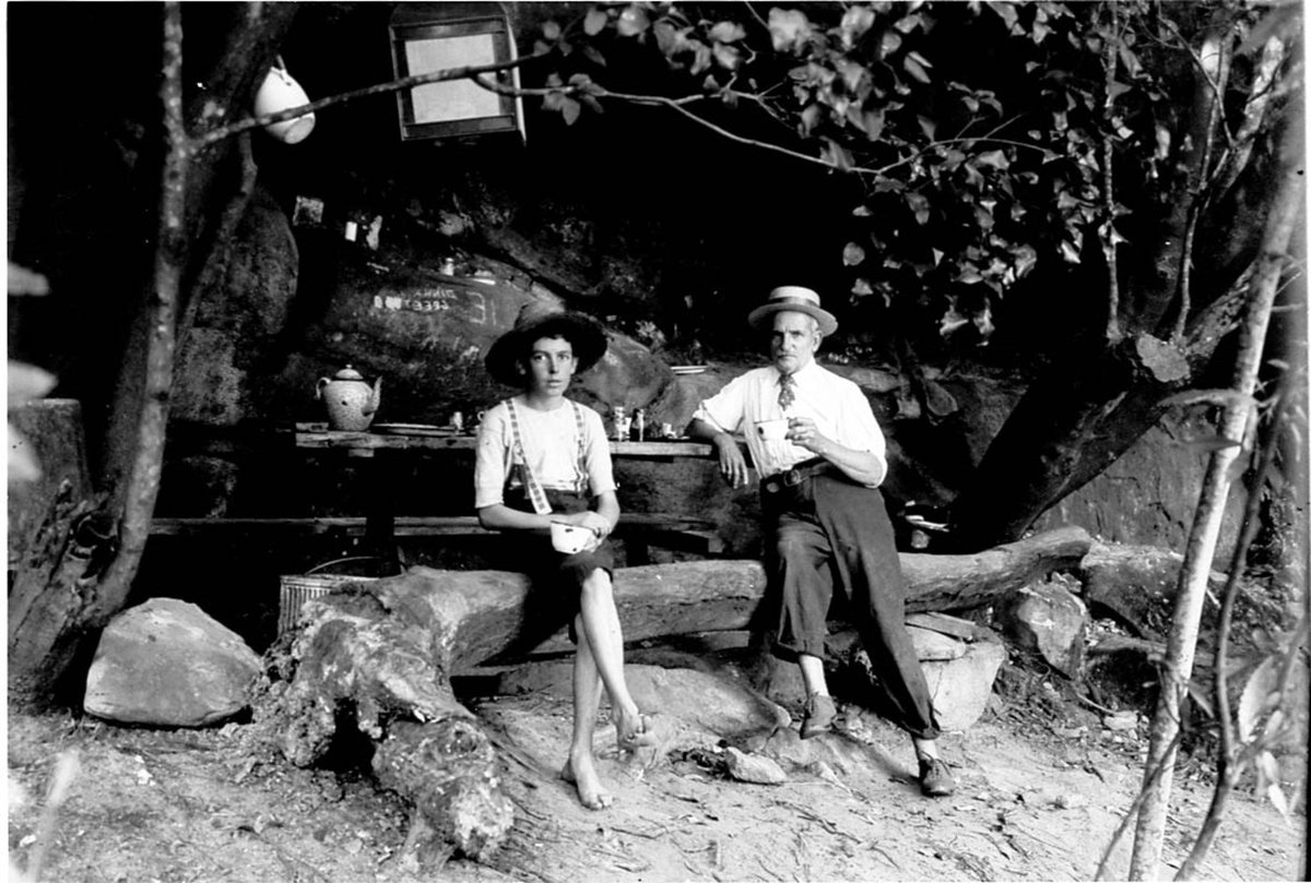 Paddy Flynn, chief clerk, Dept. of Public Works, with boy in kitchen of camp (to find site of Spit Bridge) in rock shelter, Middle Harbour, 1893, By A.J. Perier, Sydney Harbour