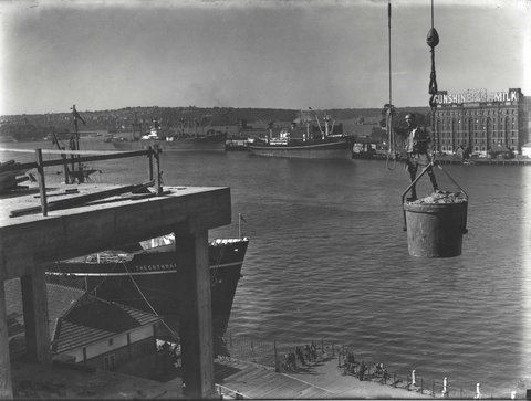 View of shipping wharves in Sydney Harbour with a construction worker standing in the bucket of a crane in the foreground, c. 1959