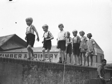 Five small boys and two girls walking along a brick wall, 1935